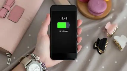 Ein schwarzes Smartphone zeigt 82 % Akkuladung in einer Hand, umgeben von einer Geldbörse, Haarspangen und Macarons.