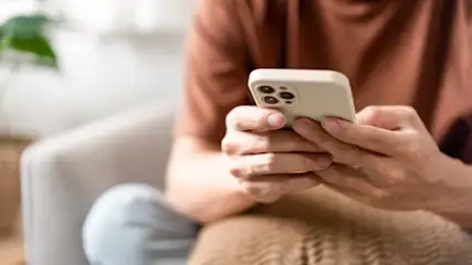 Eine Person im braunen T-Shirt sitzt und hält ein beiges Smartphone in den Händen. Ein Stuhl und eine Pflanze sind im Hintergrund.