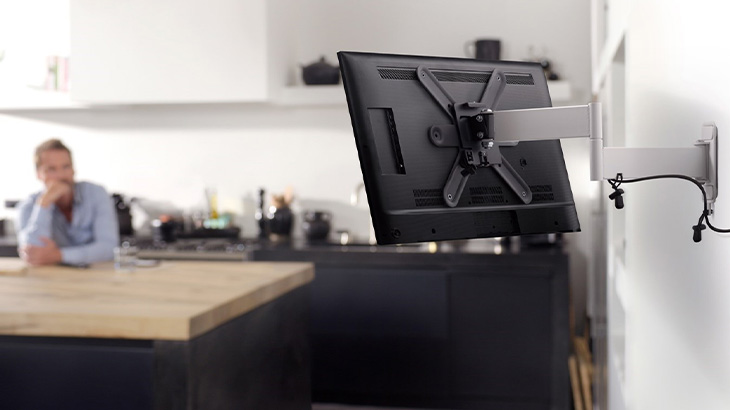 Ein an der Wand montierter Fernseher steht im Vordergrund, während eine Person an einer Küchentheke sitzt.