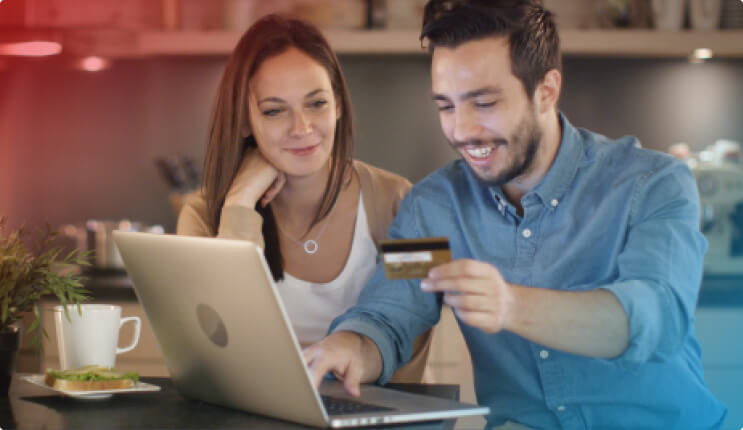 Ein Mann mit Hemd hält eine Karte vor einem Laptop, während eine Frau zusieht. Auf dem Tisch stehen eine Tasse und ein Teller.