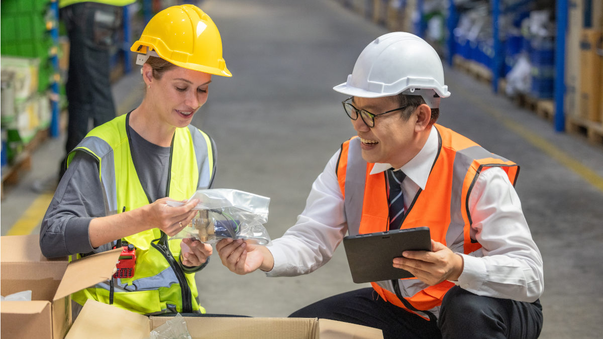 Zwei Personen mit Schutzhelmen und Sicherheitswesten inspizieren Teile in einer Lagerhalle. Einer hält ein Tablet. Regale im Hintergrund.