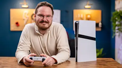 Ein Mann mit Brille hält einen Controller neben einer weißen Playstation 5 auf einem Holztisch vor einer blauen Wand.