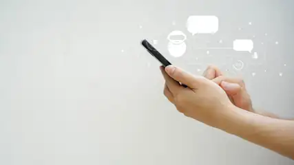 Eine Hand hält ein schwarzes Smartphone. Darüber sind Chat-Symbole und ein Roboter-Avatar auf weißem Hintergrund zu sehen.