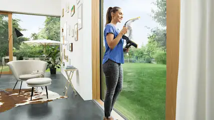 Eine Frau putzt mit einem gelben Fenstersauger ein Fenster. Draußen ist ein Garten sichtbar.