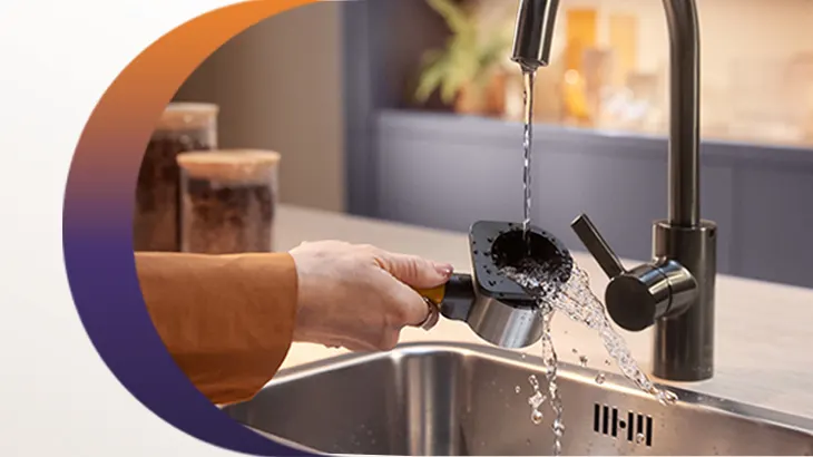 Eine Hand hält einen Siebträger unter einen Wasserhahn über einem Spülbecken. Wasser läuft aus dem Siebträger. Im Hintergrund sind Behälter.