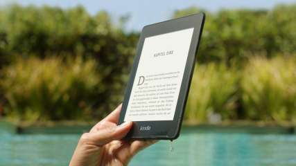 Eine Hand hält einen Kindle Ebook-Reader, im Hintergrund ist ein See zu sehen.