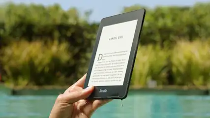 Eine Hand hält einen schwarzen Kindle mit Text auf dem Bildschirm vor einem Hintergrund aus Grün und blauem Wasser.