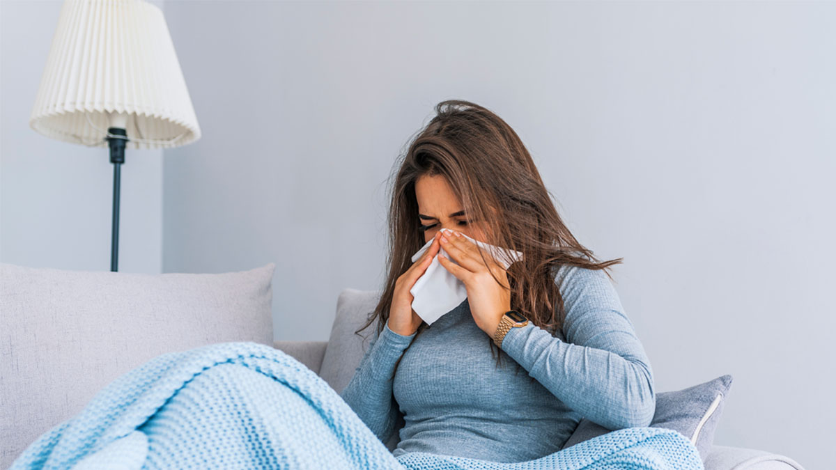 Eine Frau mit blauem Pullover auf einem Sofa hält ein Papiertuch an ihre Nase, ihre Augen sind geschlossen.