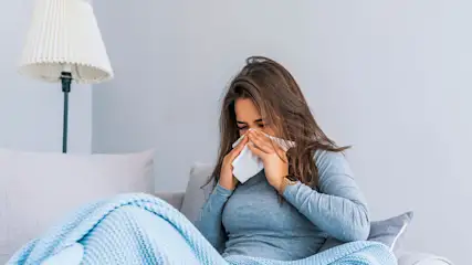 Eine Frau mit blauem Pullover auf einem Sofa hält ein Papiertuch an ihre Nase, ihre Augen sind geschlossen.