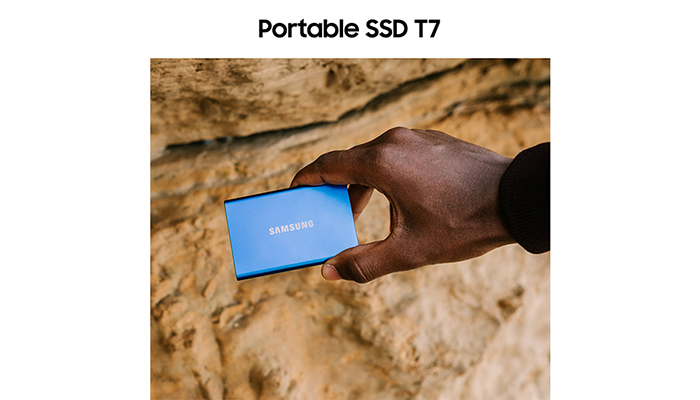 Eine Hand hält eine blaue Samsung SSD vor einem Hintergrund aus Sand.