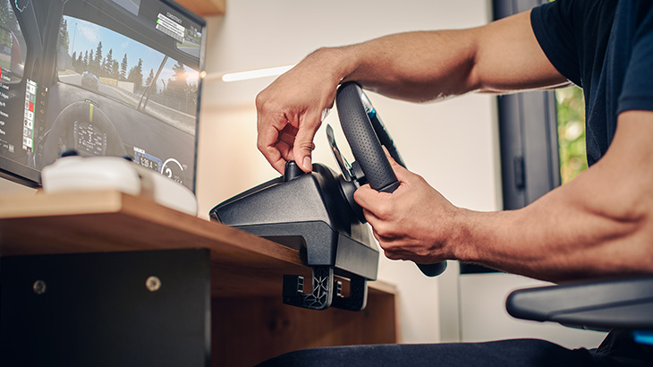 Eine Person justiert ein schwarzes Lenkrad, das an einem Holztisch befestigt ist, während ein Monitor ein Spiel zeigt.