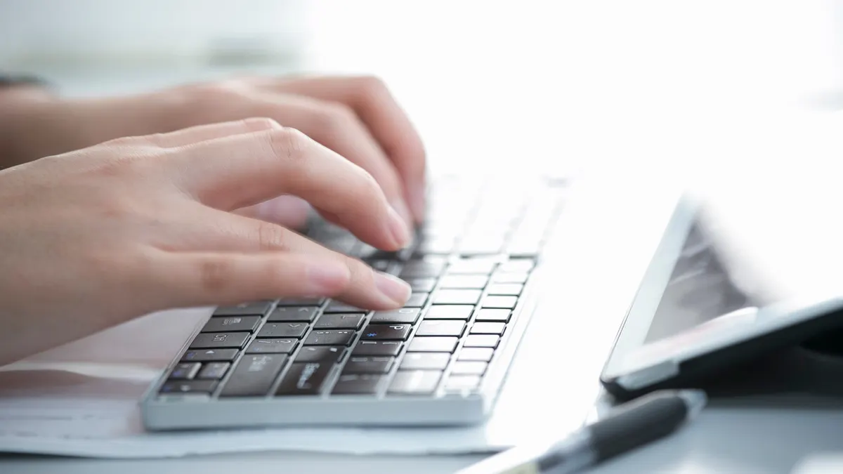 Zwei Hände tippen auf einer silbernen Tastatur, ein Tablet und ein Stift liegen daneben auf einem hellen Tisch.