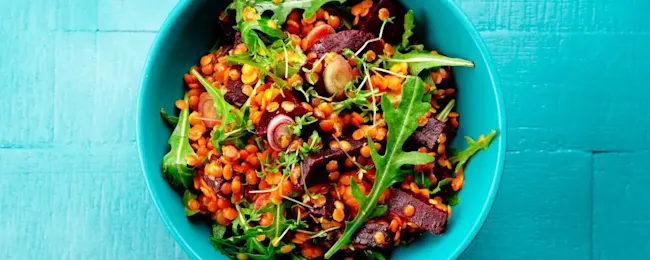 Eine Nahaufnahme einer türkisfarbenen Schüssel mit einem Salat aus braunen Linsen, grünen Blättern und roten Rüben.