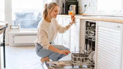 Eine blonde Frau kniet vor einer geöffneten Spülmaschine.