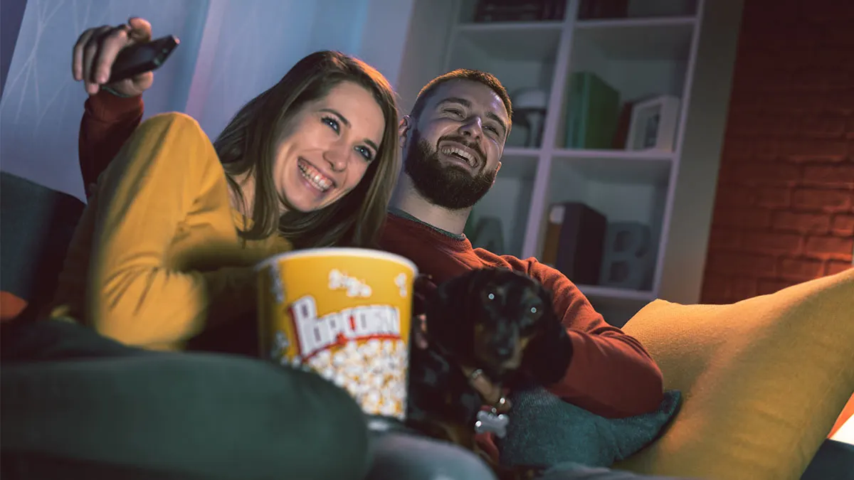 Zwei Personen und ein Hund beim Fernsehen auf der Couch. Im Vordergrund ein Popcorneimer.