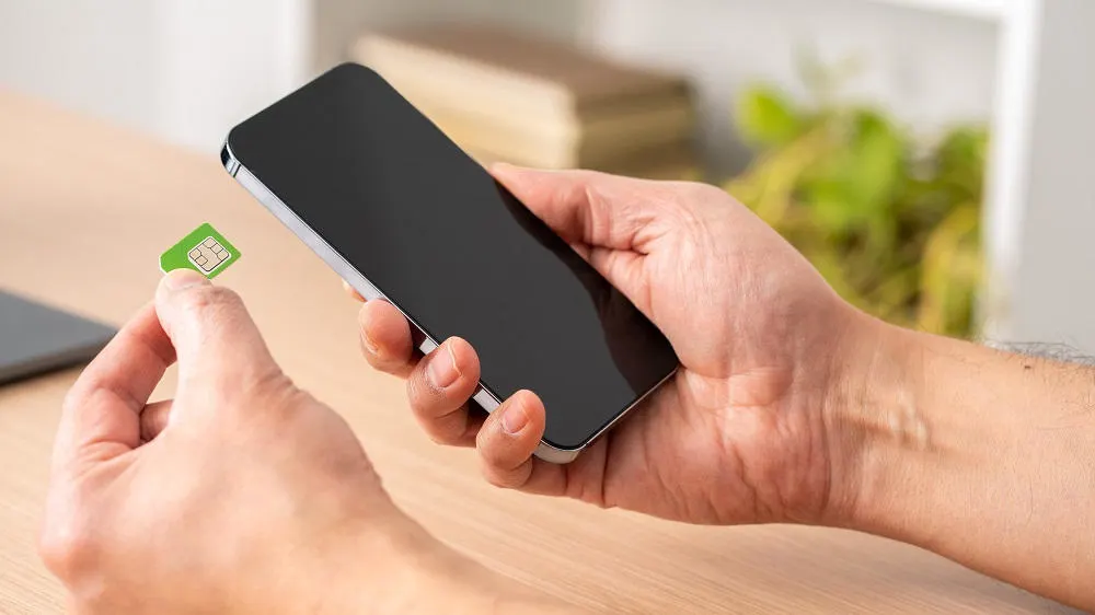 Eine Hand hält ein schwarzes Smartphone, während die andere eine grüne SIM-Karte in der Nähe auf einem Holztisch hält.