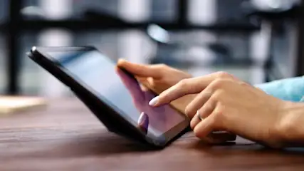 Zwei Hände tippen auf einem schwarzen Tablet auf einem braunen Tisch. Im Hintergrund sind Säulen erkennbar.