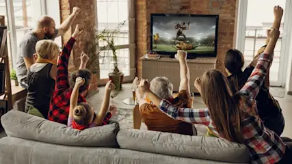 Eine Gruppe von Leuten, die auf einem Sofa sitzt und Football im Fernsehen schaut, jubelt mit erhobenen Armen.