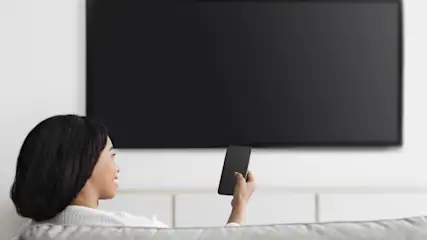 Eine Frau sitzt auf einem Sofa mit einem schwarzen Smartphone in der Hand vor einem ausgeschalteten Fernseher.