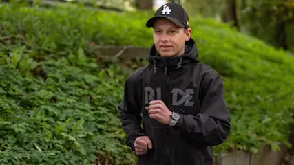 Ein Mann mit schwarzer Jacke und Kappe joggt vor grünem Laub. Er trägt eine Uhr.