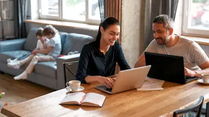 Eltern arbeiten an Laptops am Holztisch. Kinder sitzen auf einem grauen Sofa im Hintergrund. Kaffeetassen stehen auf dem Tisch.