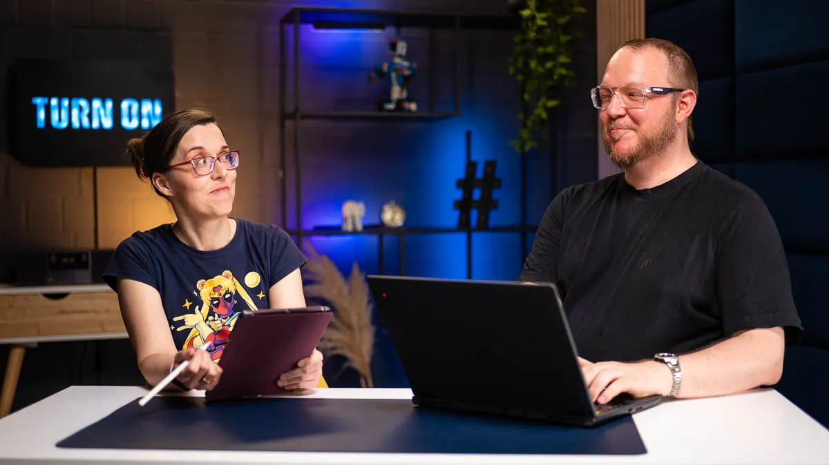 Daniela sitzt mit ihrem iPad neben Andreas und seinem Laptop an einem Tisch und schaut Andreas ungläubig an.