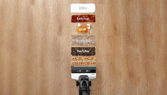 Ein Staubsauger auf einem Holzboden saugt Flecken wie Asche, Ketchup, Ei, Tierhaare, Tierfutter und groben Schmutz auf.