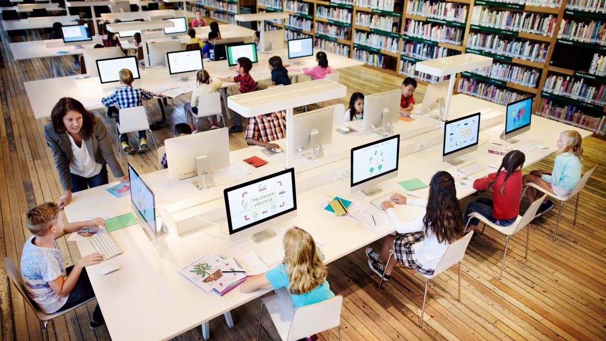 SchülerInnen lernen an iMacs in einer Bibliothek unter Lehraufsicht.