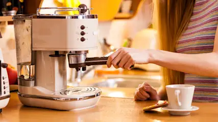 Eine Person bereitet in einer hellen Küche einen Kaffee an einer Kaffeemaschine zu.