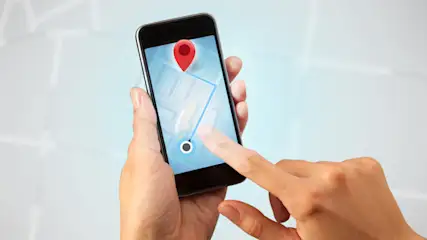 Eine Hand hält ein Smartphone mit einer Karten-App, auf der ein roter Marker und eine blaue Route zu sehen sind.