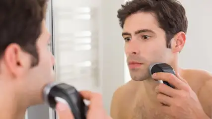 Ein Mann rasiert sich mit einem elektrischen Rasierer vor einem Spiegel im Badezimmer.