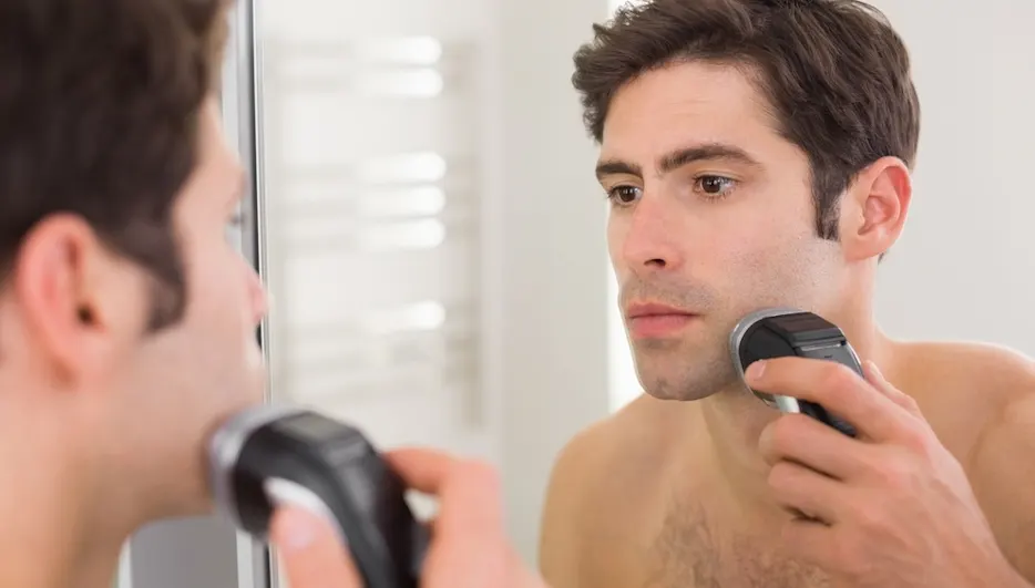Ein Mann rasiert sich mit einem elektrischen Rasierer vor einem Spiegel im Badezimmer.