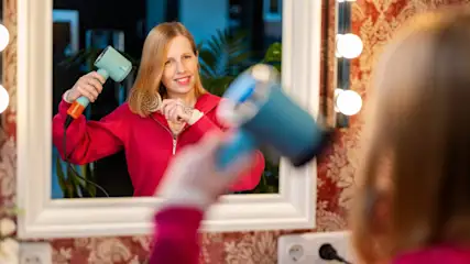 Eine Frau hält einen Föhn und eine Bürste vor einem Spiegel mit Glühbirnen und floraler Tapete.