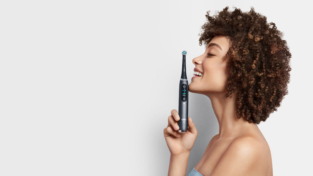 Eine lächelnde Frau hält eine elektrische Zahnbürste von Oral-B hoch.