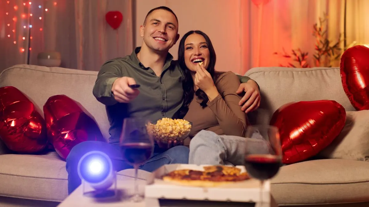 Ein Paar sitzt mit Popcorn auf dem Sofa. Vor den beiden steht ein eingeschalteter Mini-Beamer auf einem Tisch.