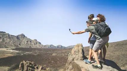 Eine Gruppe Wanderer steht auf einem Felsen mit Blick auf eine Berglandschaft unter blauem Himmel. Ein Mann hält eine Kamera.
