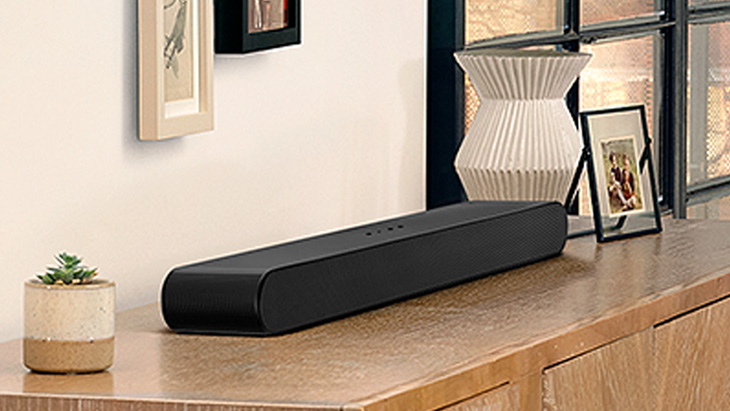 Schwarzer Lautsprecher auf einem Holzschrank, neben einer Topfpflanze, einer Vase und gerahmten Bildern.