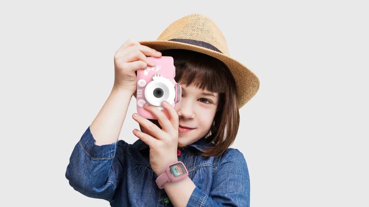 Ein Kind mit Hut und Jeanshemd hält eine rosa Kamera vor dem Gesicht und trägt eine rosa Uhr.