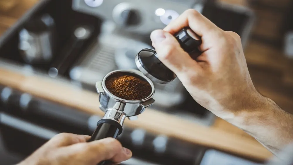 Zwei Hände halten einen Siebträger mit Kaffeepulver und einen Tamper vor einer Kaffeemaschine.