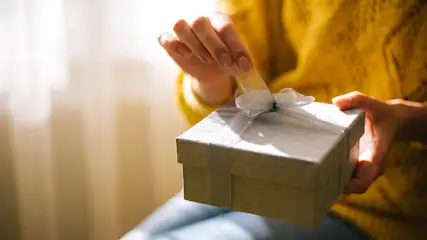 Eine Person in gelbem Pullover öffnet eine quadratische, weiße Geschenkbox mit Schleife.