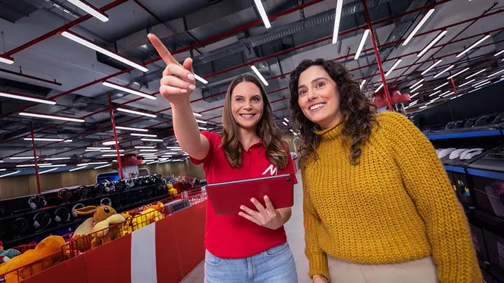Zwei Frauen stehen in einem Lagerhaus; eine zeigt mit dem Finger nach oben, die andere lächelt und trägt einen gelben Pullover.