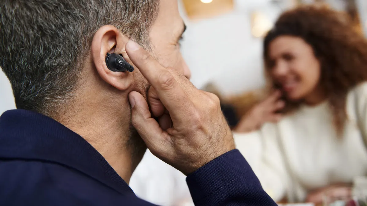 Ein Mann mit einem schwarzen Ohrhörer im Ohr berührt ihn mit dem Finger. Im Hintergrund ist eine Frau zu sehen.