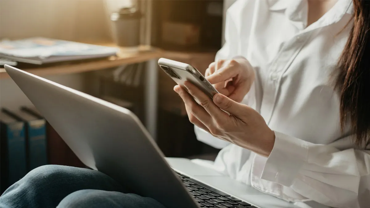 Eine Frau auf einem Sofa hält ein Smartphone in der Hand und einen Laptop auf dem Schoß.