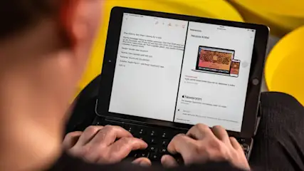 Eine Person tippt auf einem schwarzen Tablet mit Tastatur. Der Bildschirm zeigt geteilten Text und ein Newsroom-Layout vor gelbem Hintergrund.