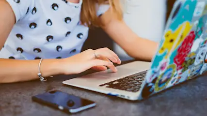 Eine Person tippt auf einem Laptop mit Aufklebern. Ein Smartphone liegt daneben auf einem grauen Tisch. Person trägt Armband und Ring.