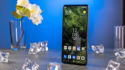 Ein Smartphone mit grünem Bildschirm liegt auf einem Tisch, umgeben von Eiswürfeln. Eine Vase mit weißen Blüten steht daneben.