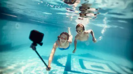 Zwei Kinder mit Schwimmbrillen schwimmen in einem Pool und filmen sich mit einer Kamera am Selfie-Stick.