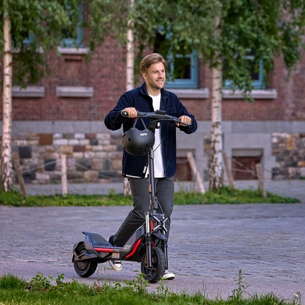 Ein Mann steht auf einem grauen Roller vor einem Gebäude mit Bäumen. Er hält einen schwarzen Helm.