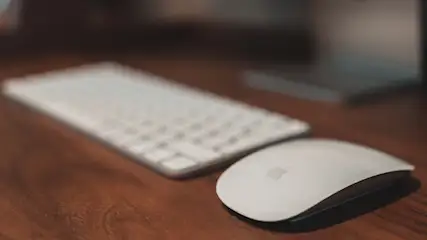 Eine weiße Computermaus liegt neben einer weißen Tastatur auf einem braunen Holztisch.