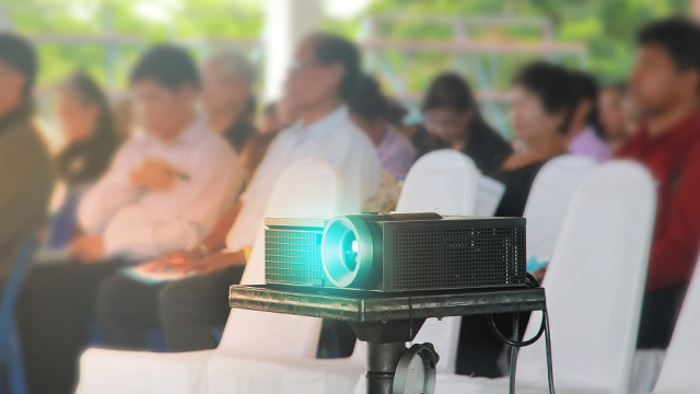 Ein Projektor steht auf einem Tisch, im Hintergrund sitzen Personen auf weißen Stühlen. Blaue Lichtstrahlen kommen aus dem Projektor.